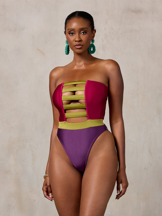 A woman modeling a strapless one-piece swimsuit with a magenta-pink bodice, olive green horizontal stripes across the chest, and a deep purple high-cut bottom, standing against a textured beige studio wall. MBM Swim by Marcia B Maxwell colorful zen one-piece swimsuit on model #color_tropic-summer