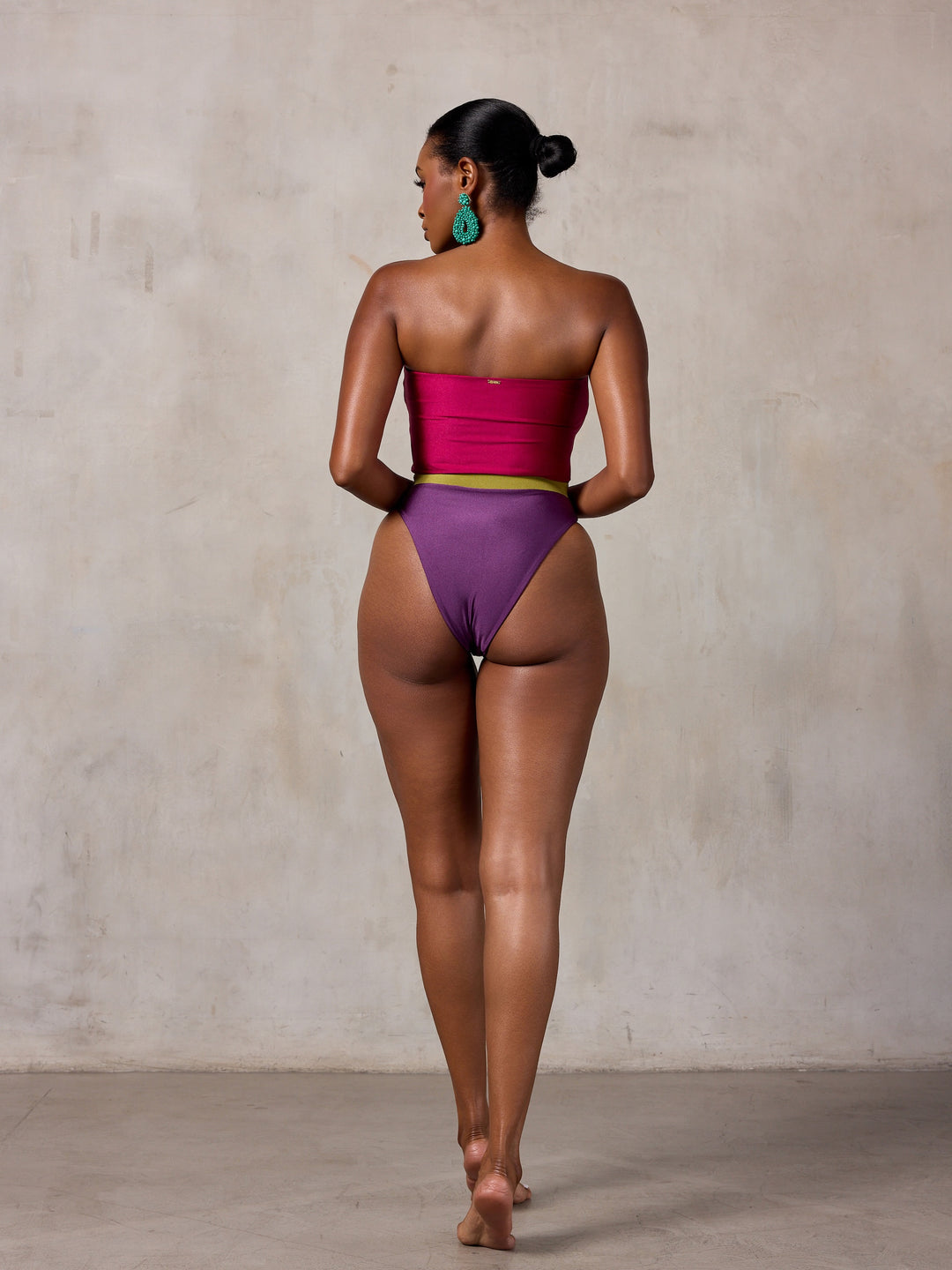 A woman modeling a strapless one-piece swimsuit with a magenta-pink bodice, olive green horizontal stripes across the chest, and a deep purple high-cut bottom, standing against a textured beige studio wall. MBM Swim by Marcia B Maxwell colorful zen one-piece swimsuit on model #color_tropic-summer