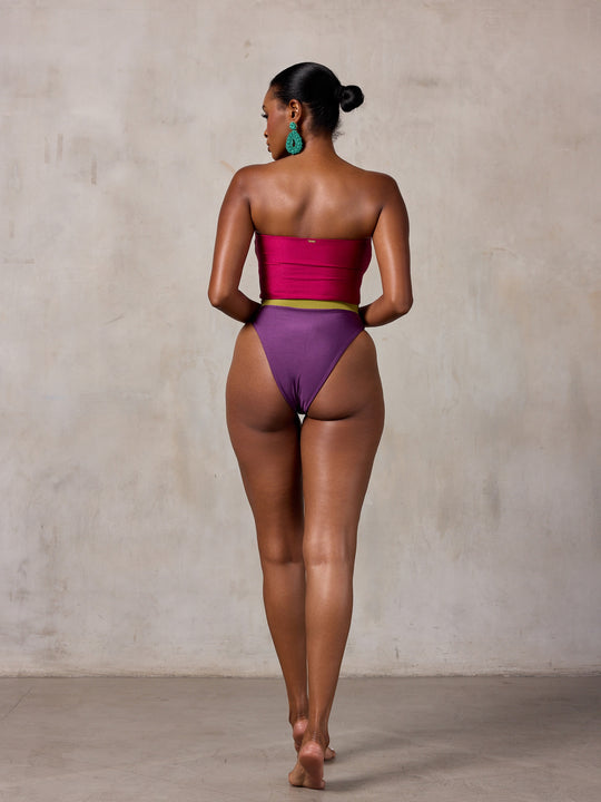 A woman modeling a strapless one-piece swimsuit with a magenta-pink bodice, olive green horizontal stripes across the chest, and a deep purple high-cut bottom, standing against a textured beige studio wall. MBM Swim by Marcia B Maxwell colorful zen one-piece swimsuit on model #color_tropic-summer